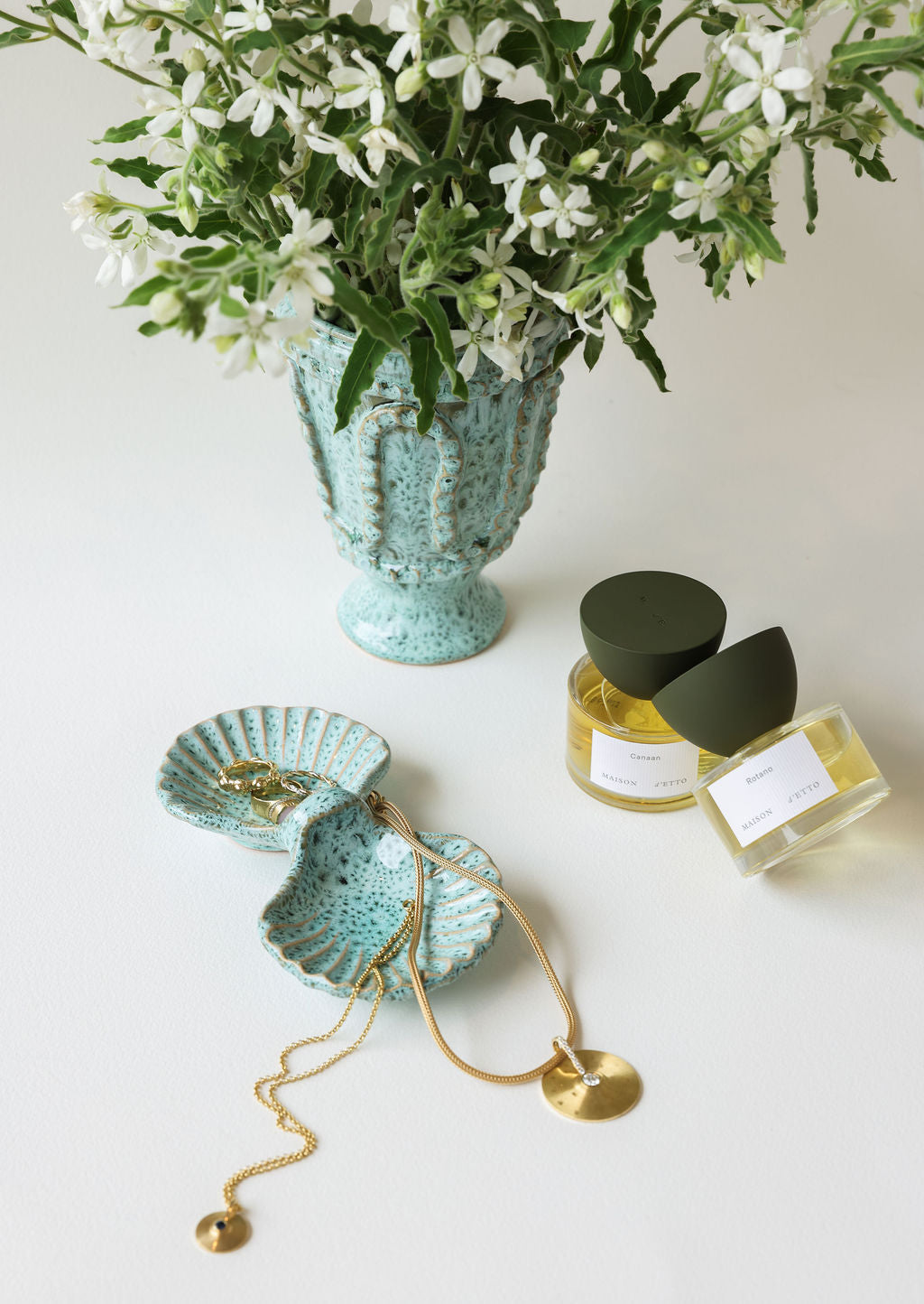 Teal ceramic vase with white flowers, green shell jewelry dish, gold necklace and diamond pendant, and two perfume bottles on a light background